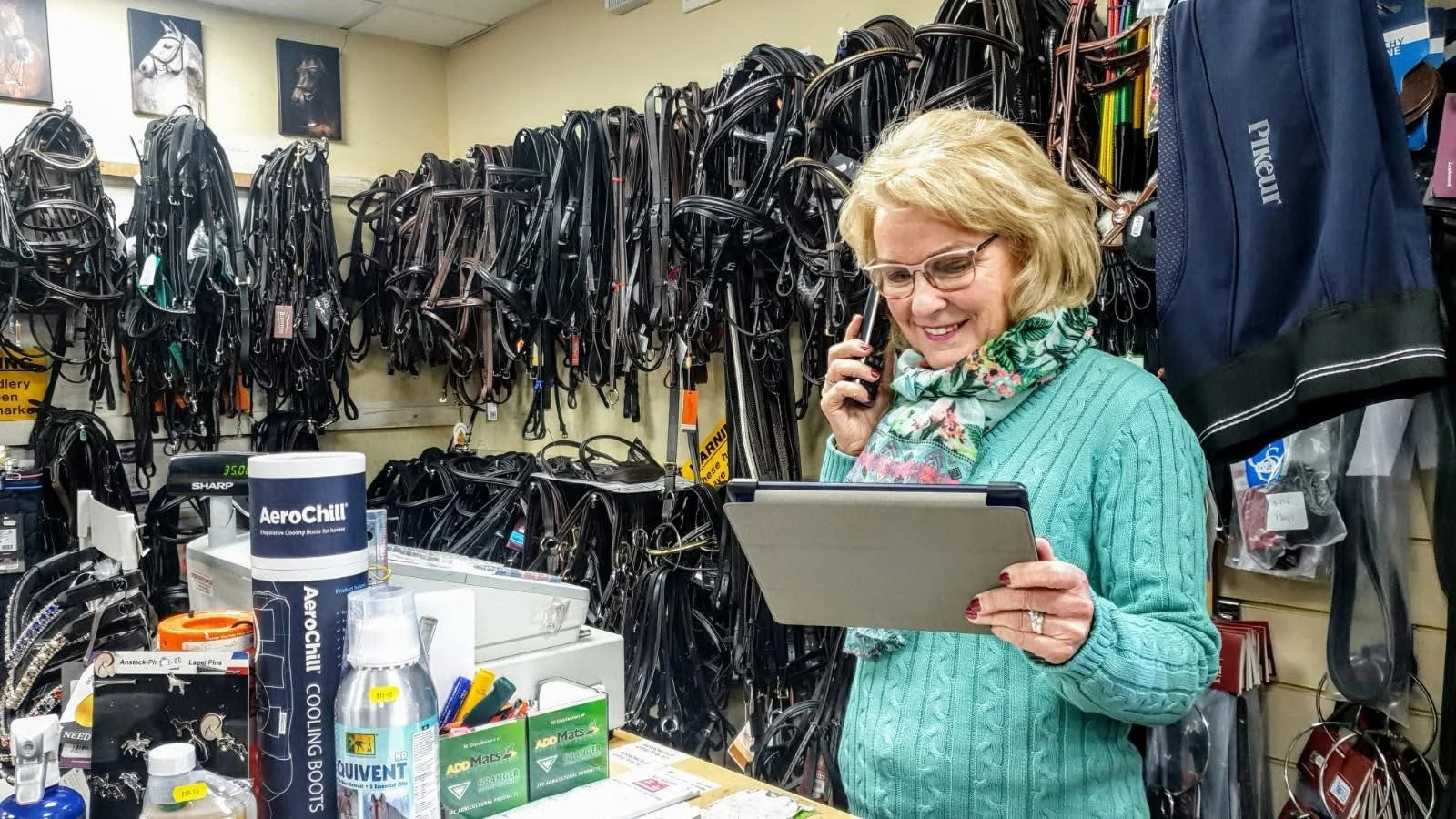 A woman in a teal sweater and floral scarf, wearing glasses, smiling while talking on a phone in a tack shop. She holds a clipboard or tablet. The background features a wall filled with horse bridles and bits, and the counter displays equine care and shoeing products.