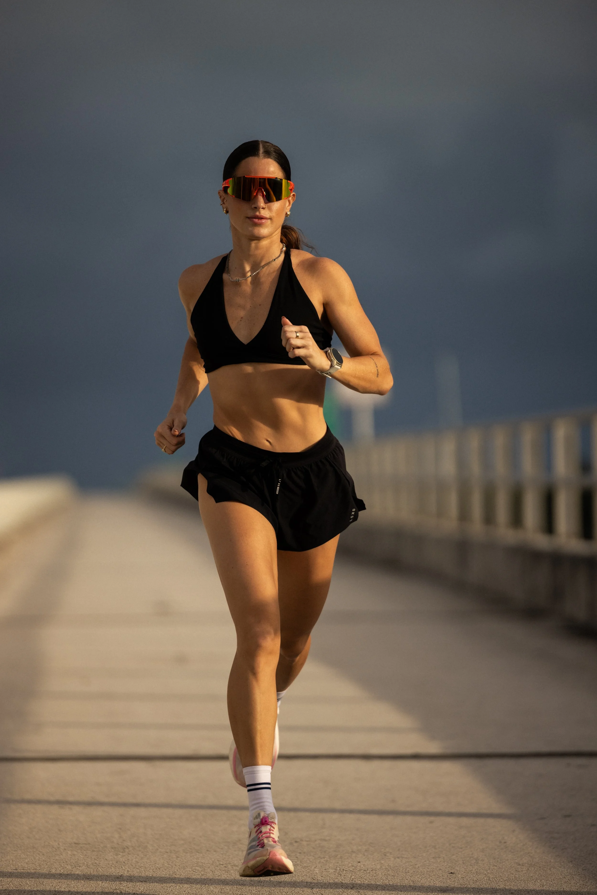 A woman running on a bridge during dusk. She is wearing a black sports bra, black shorts, white athletic shoes with pink accents, sunglasses, and a watch, with stormy clouds in the background.