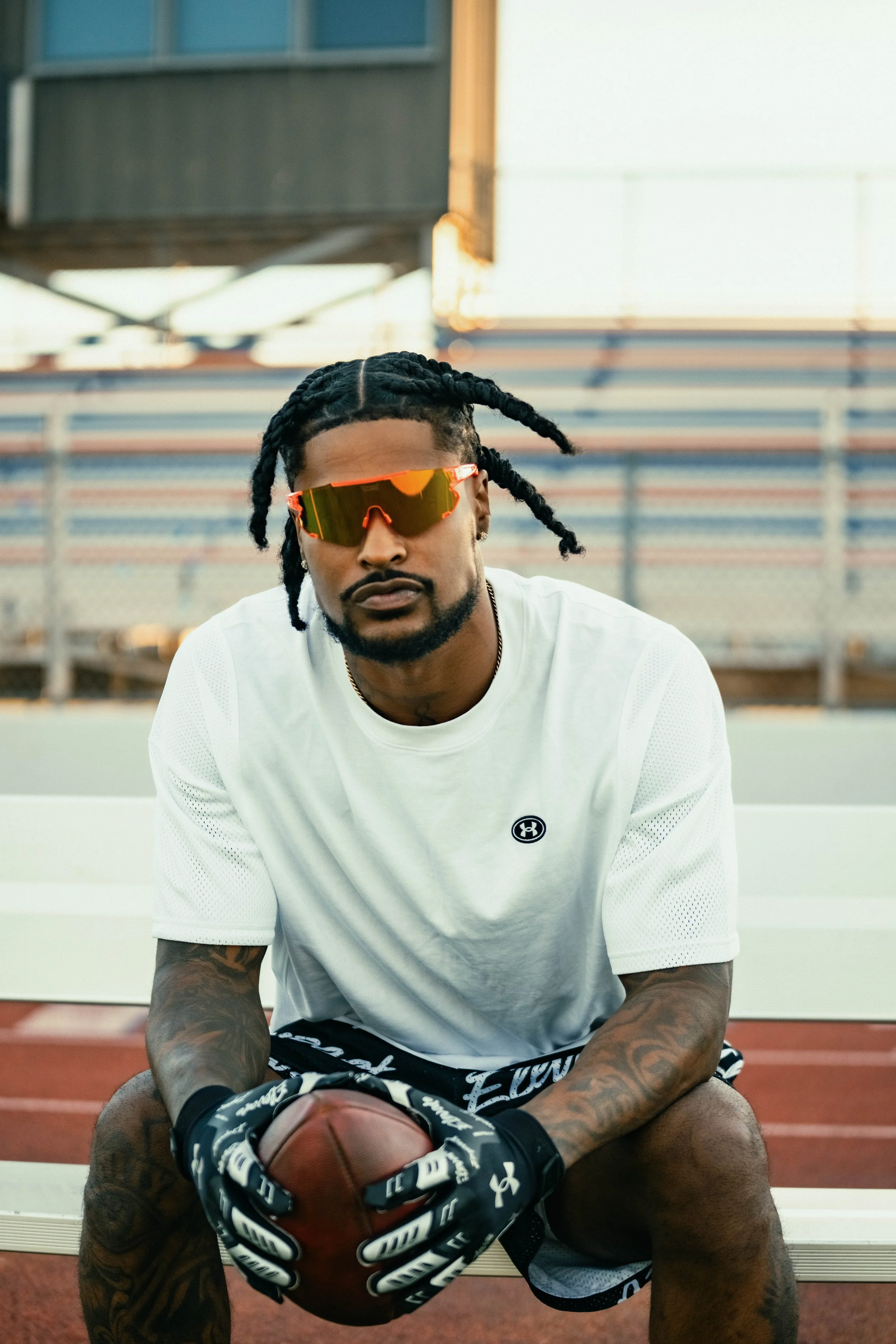 A man sitting on a white bench on an outdoor athletic track, holding a football, wearing sunglasses, a white athletic shirt, and black gloves.