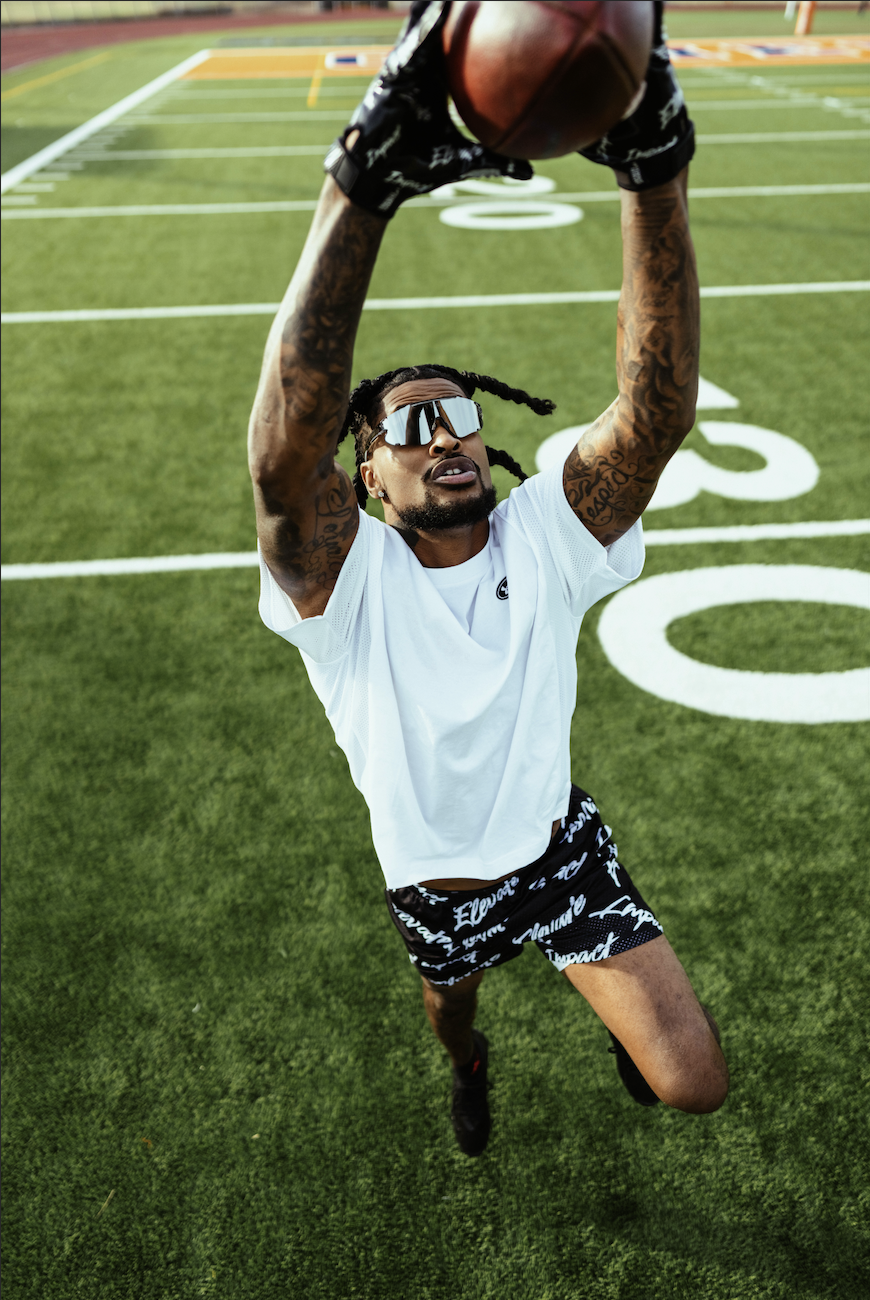 A man with tattoos and sunglasses jumping in the air on a football field, reaching up with both hands to catch a football.