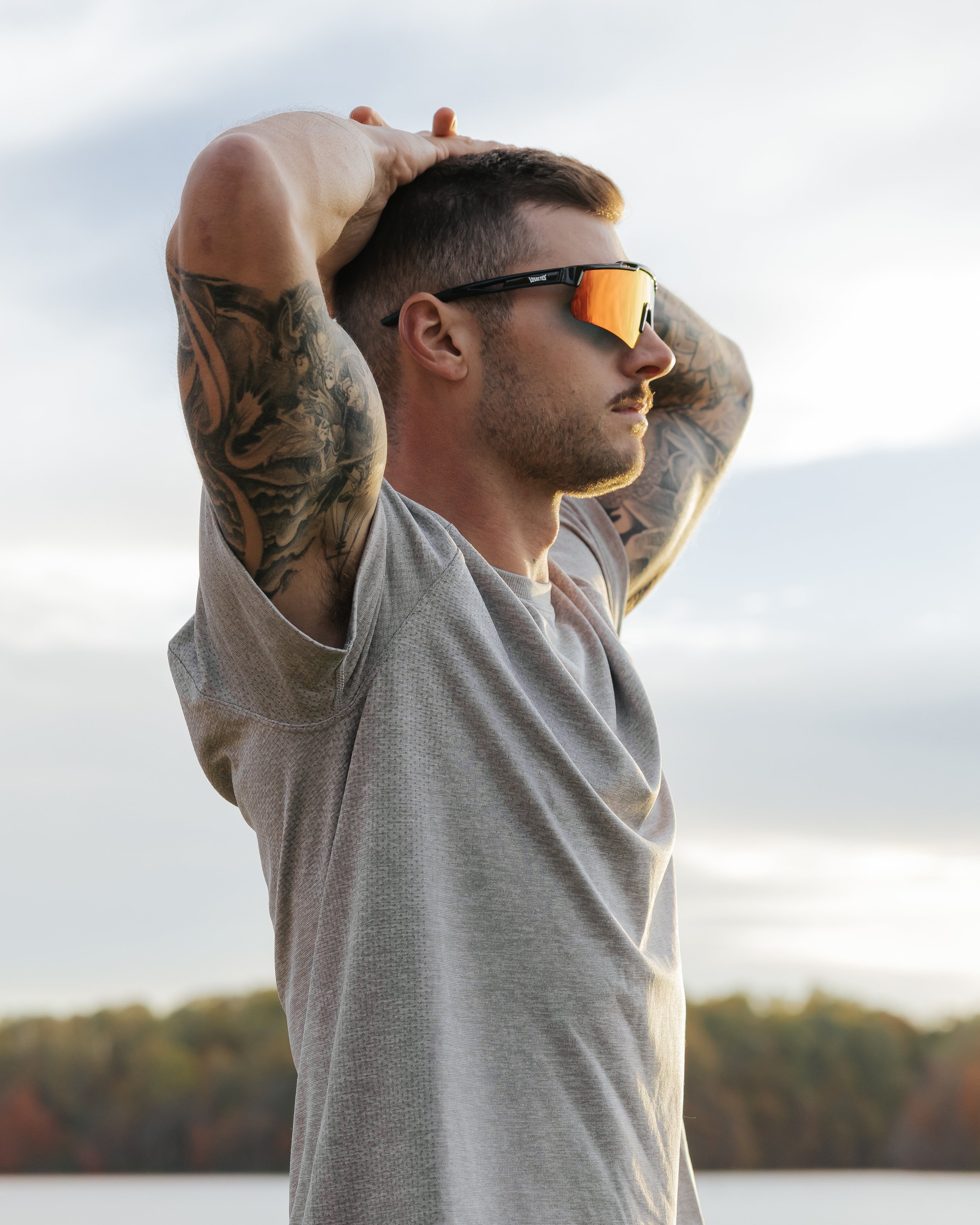 A man with tattoos on his arms wearing orange reflective sunglasses and a gray athletic shirt, standing outdoors with a lake and trees in the background, stretching with his hands behind his head.