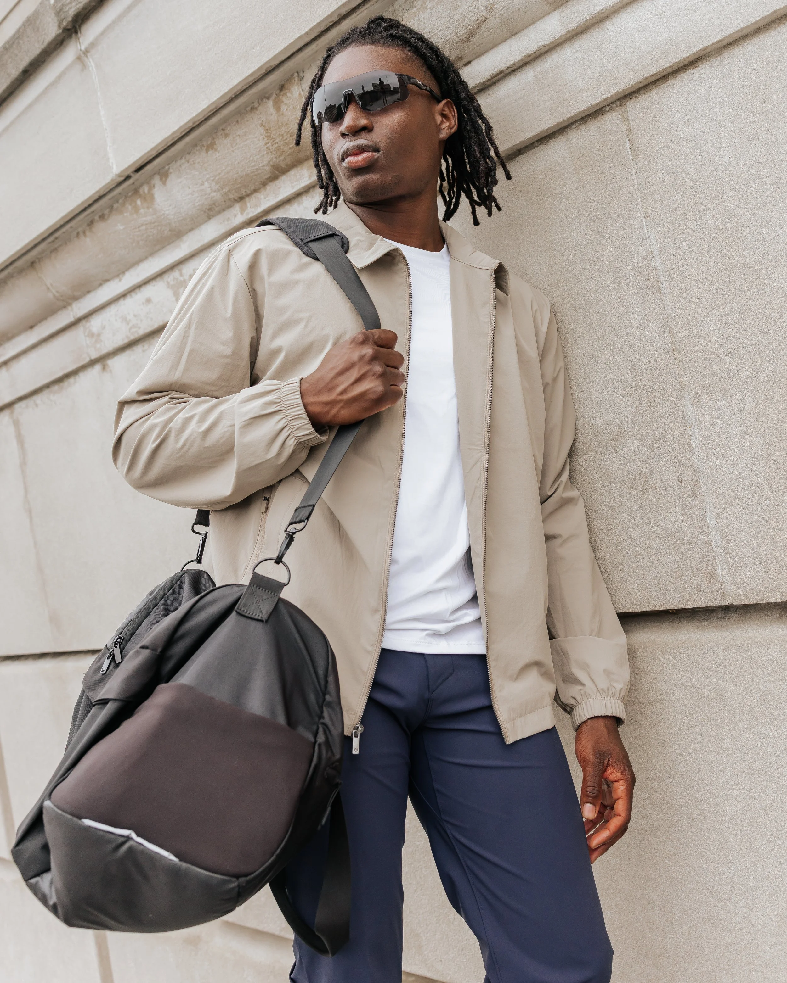 A young man with dreadlocks wearing sunglasses, a beige windbreaker, white T-shirt, navy blue pants, and carrying a black backpack with his right hand, standing against a stone wall.