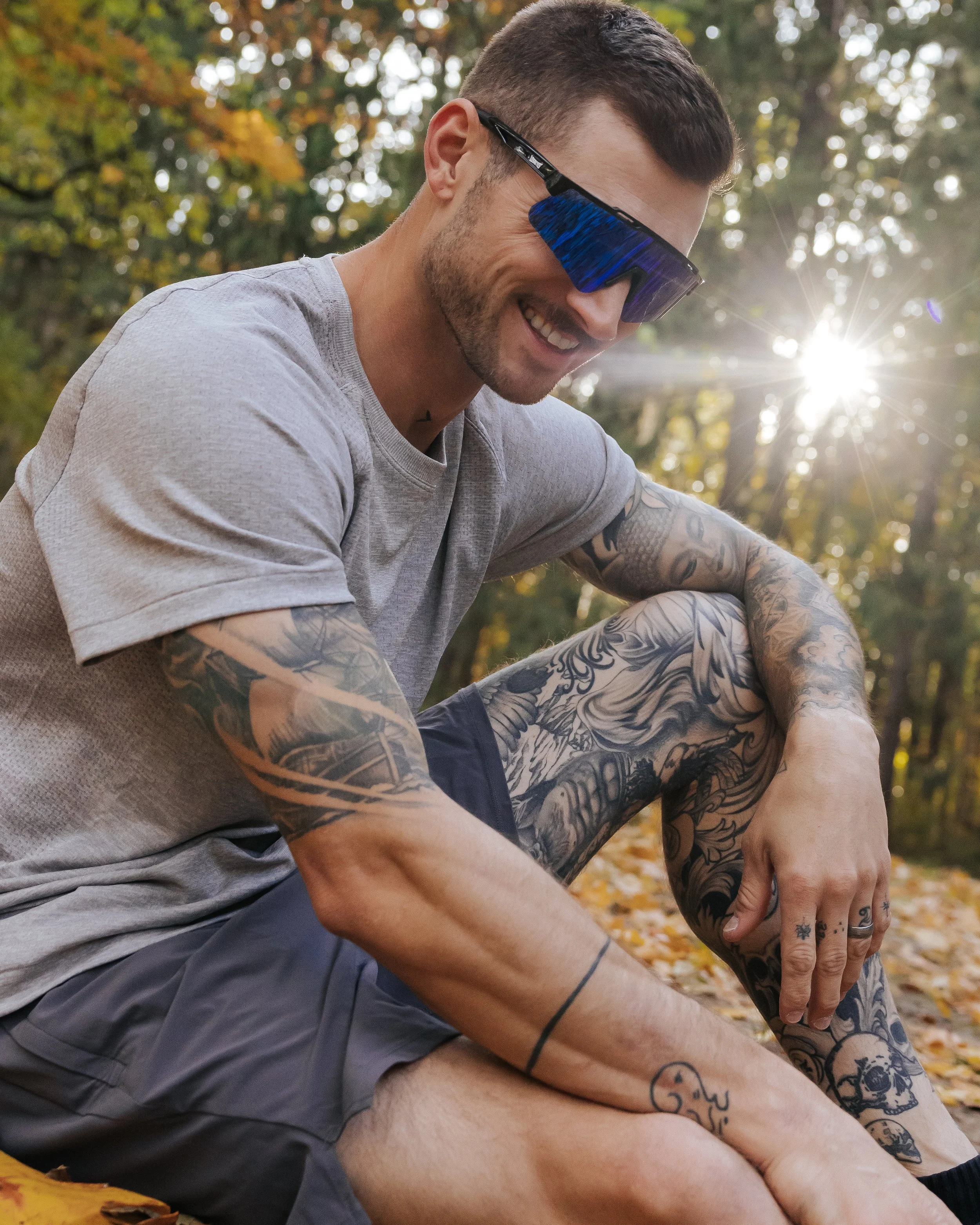 A smiling man with tattoos wearing sunglasses and a gray t-shirt sitting outdoors in a wooded area with fallen leaves, bathed in sunlight.