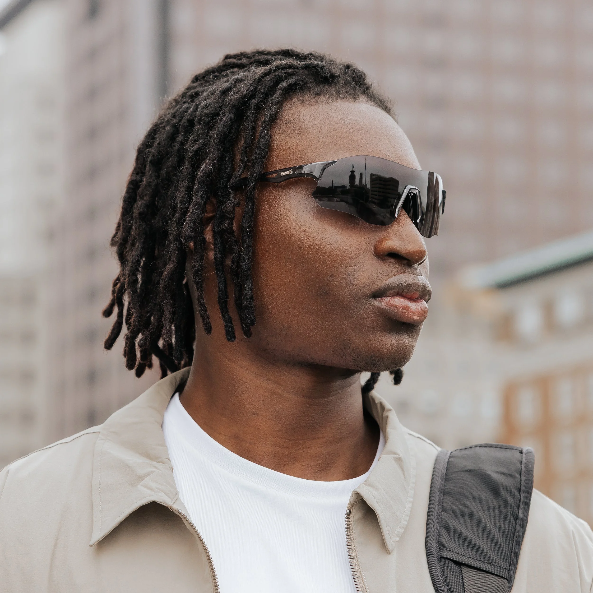 A man with dark skin and dreadlocks wearing black sports sunglasses, a beige jacket, and a white shirt, standing outdoors in an urban setting.