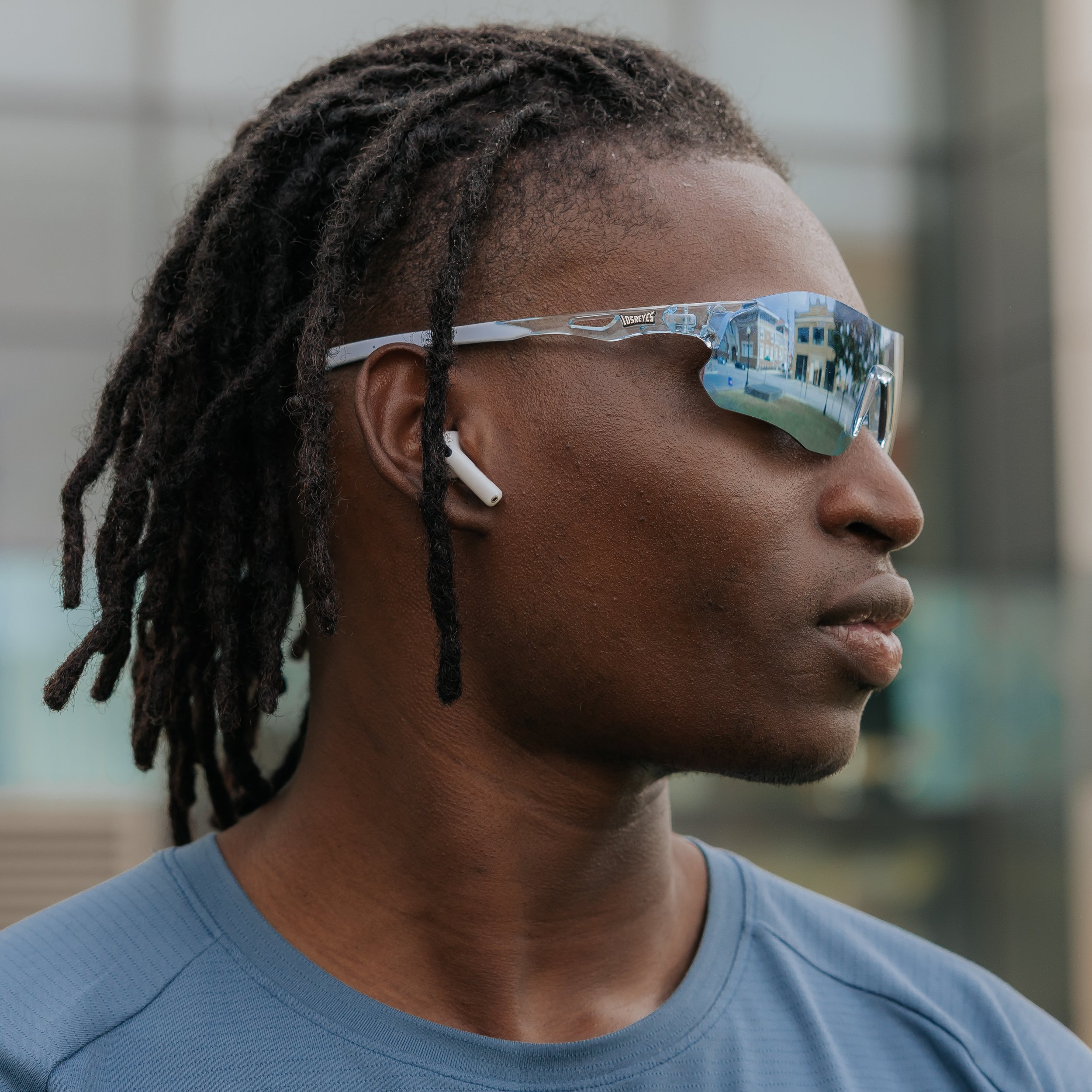 A man with dark skin and dreadlocks wearing sports sunglasses and wireless earbuds, wearing a blue athletic shirt, outdoors.
