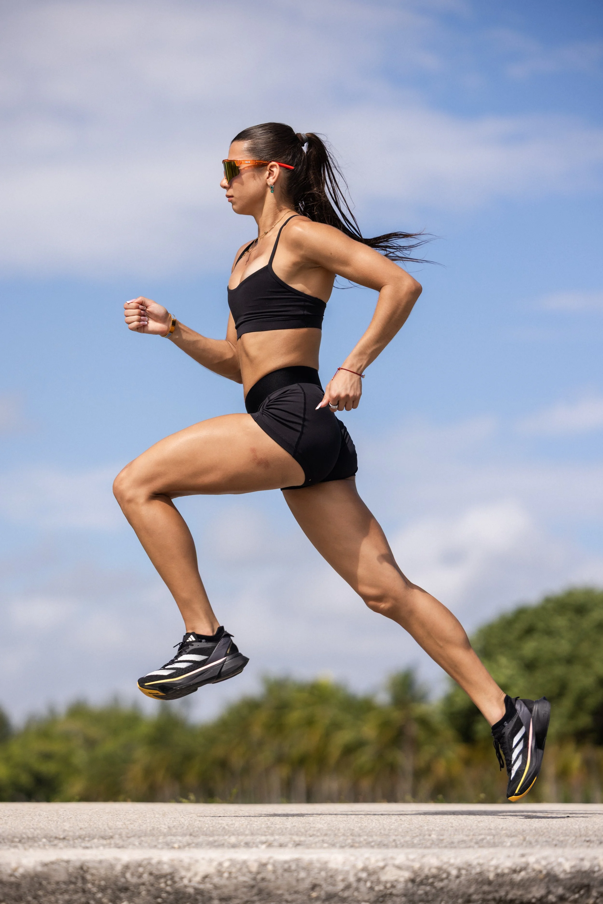 A woman running outdoors in athletic gear, wearing sunglasses, a black sports top, shorts, and running shoes, under a blue sky with some clouds and greenery in the background.