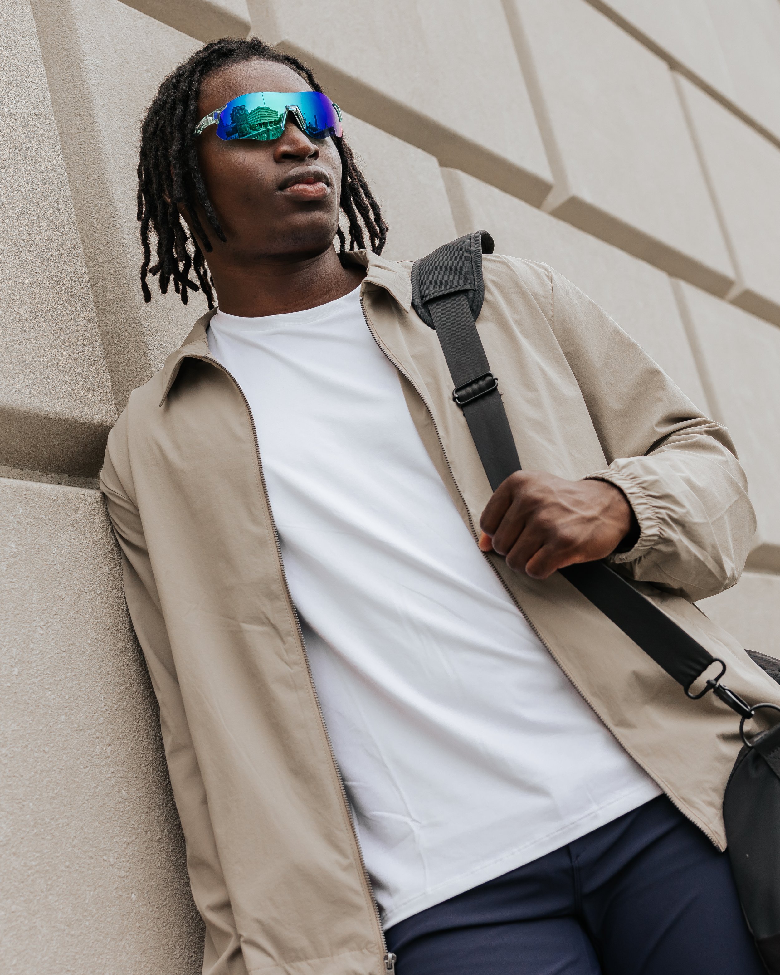 Young man with dreadlocks wearing reflective sunglasses, a beige jacket, white t-shirt, dark pants, and carrying a black backpack, leaning against a beige stone wall.