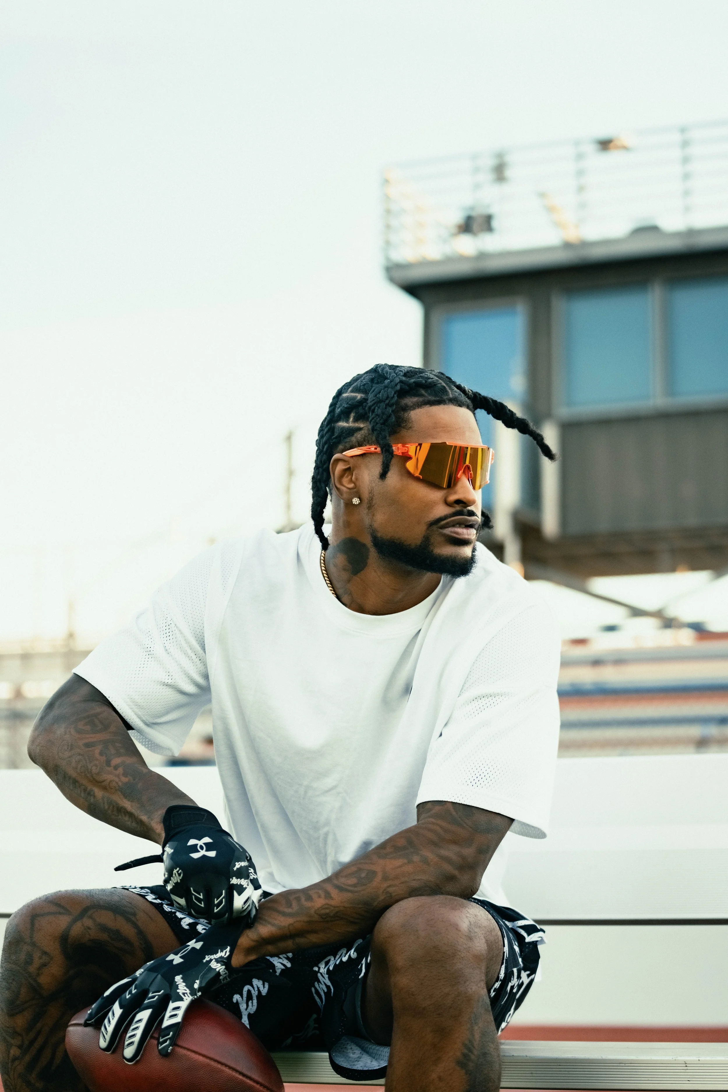 A man with tattoos, wearing sunglasses, a white athletic shirt, and gloves, sitting on a football at an outdoor sports facility.