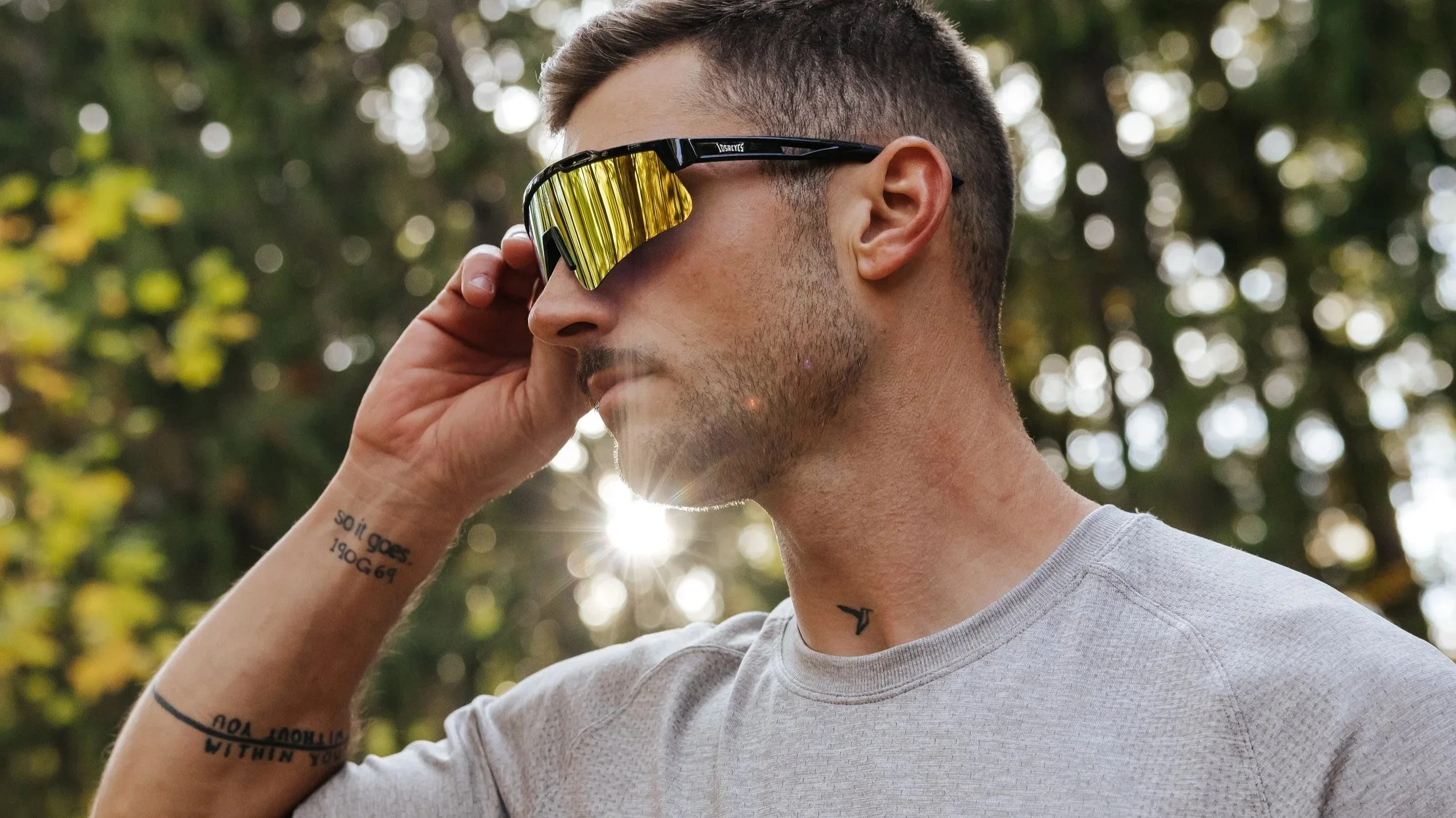 A young man with tattoos on his arm and neck wearing reflective sports sunglasses and a light gray shirt, outdoors with sunlight filtering through trees in the background.