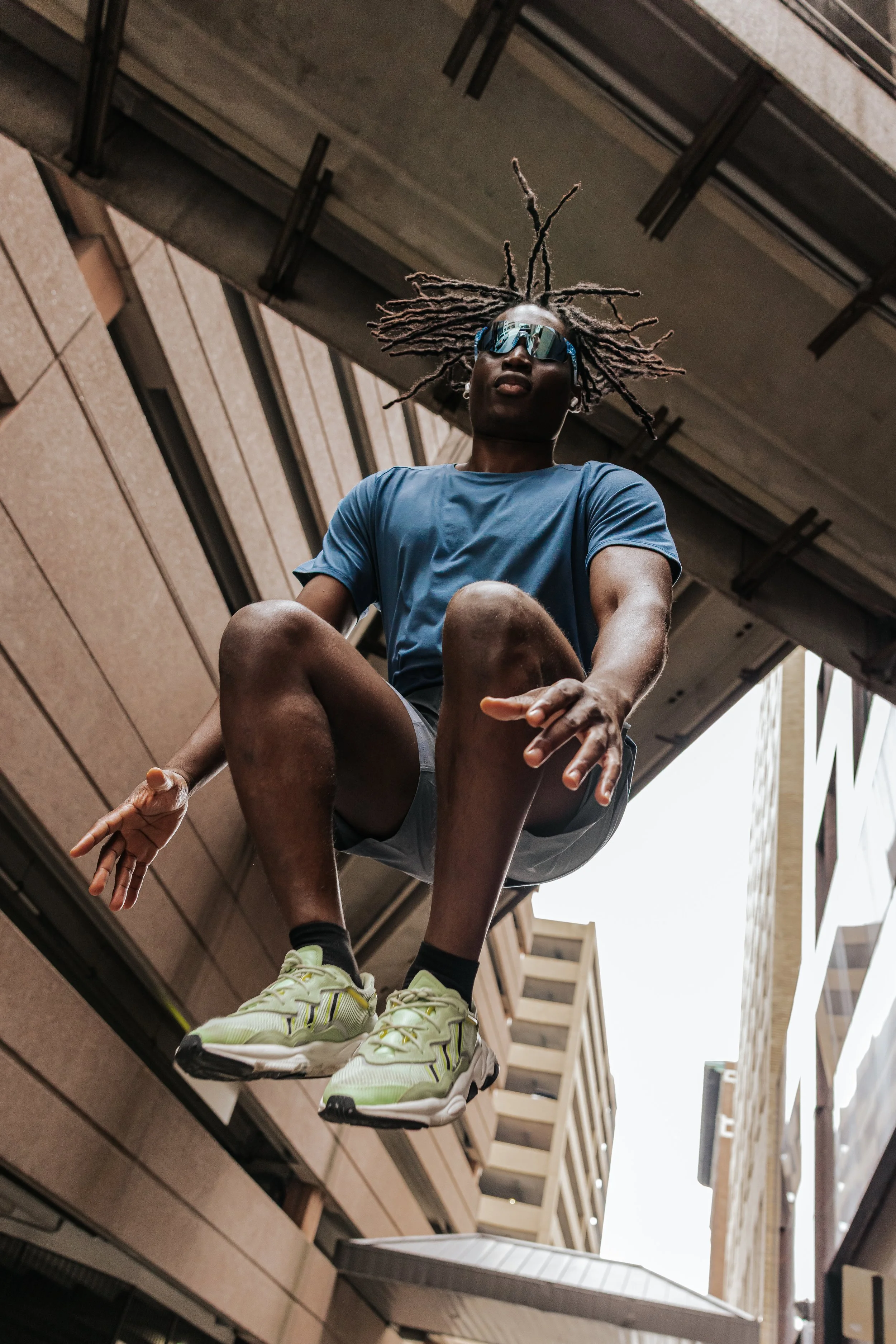 Person mid-air during parkour jump between city buildings, wearing sunglasses, blue t-shirt, shorts, and running shoes, image taken from below.