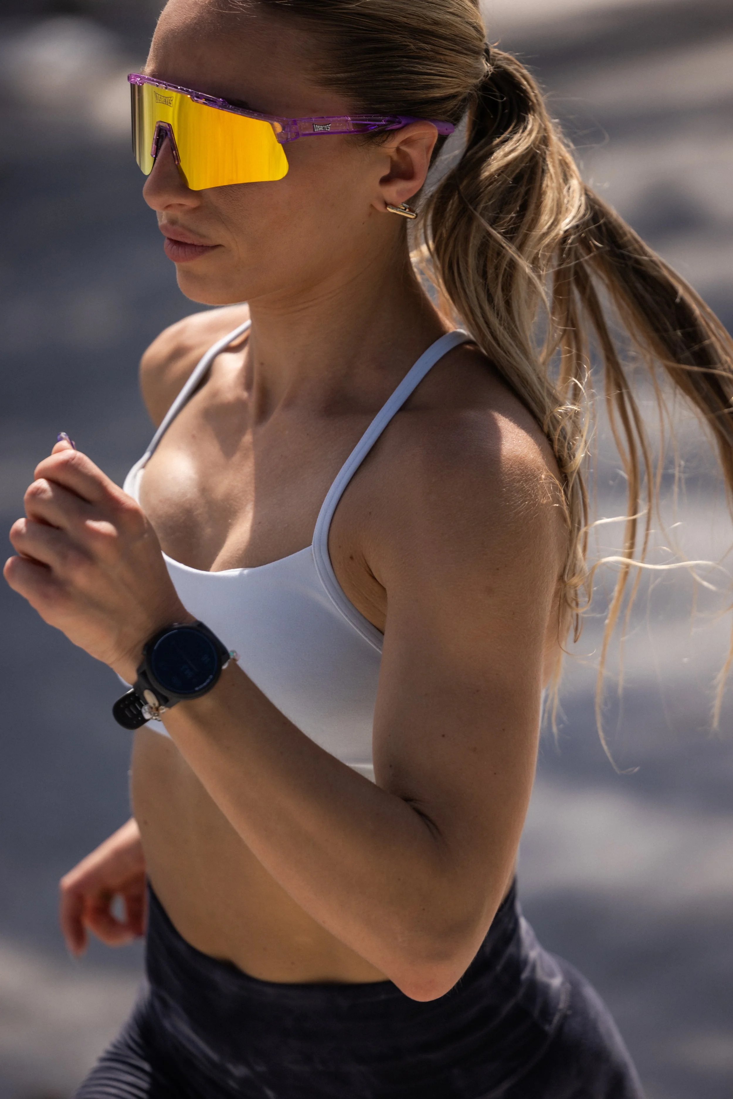 A woman running outdoors wearing sunglasses, a white sports bra, and a smartwatch, with her blonde hair in a ponytail.