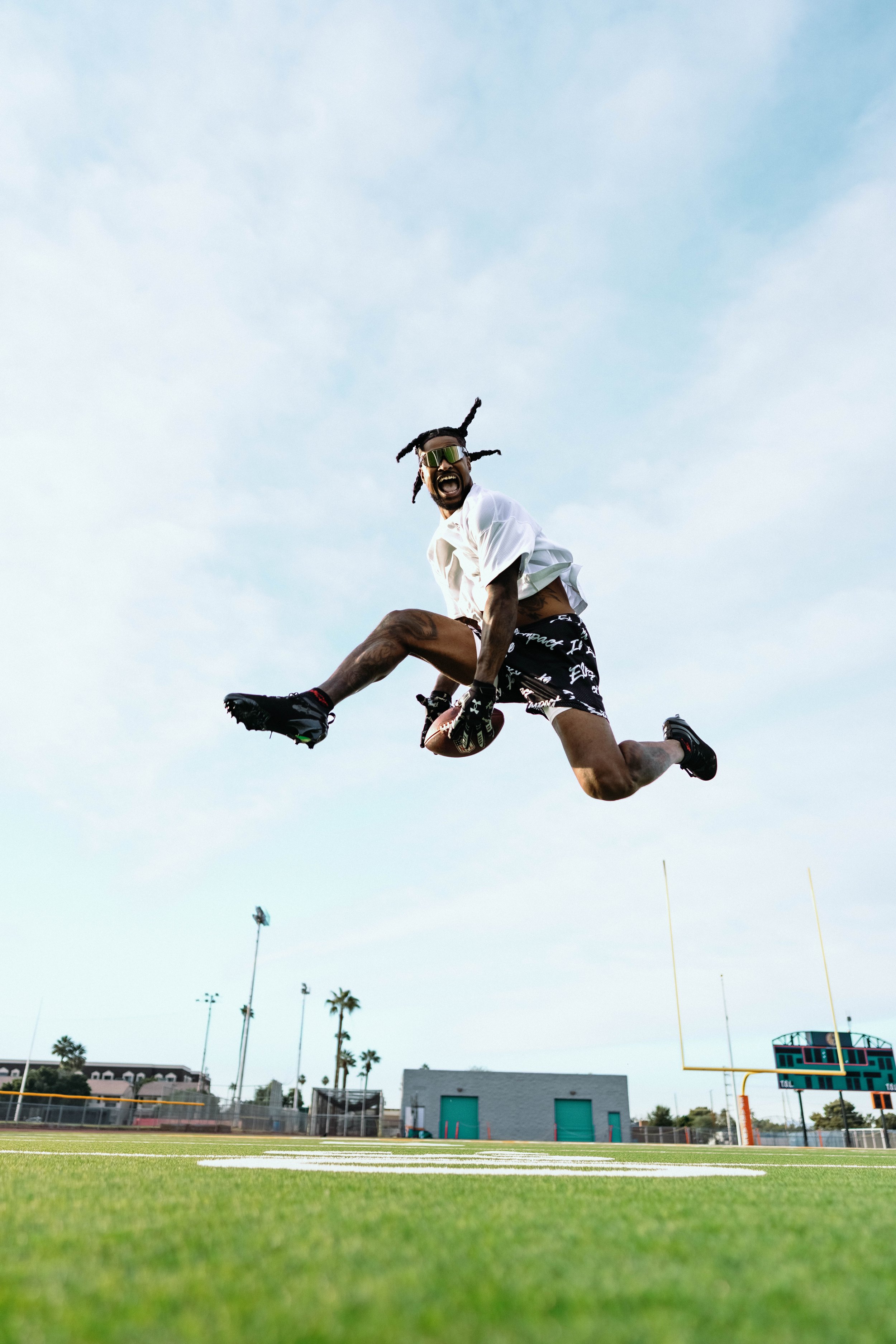 An athlete jumping in the air on a football field holding a football, wearing black shorts, a white shirt, black shoes, and sunglasses, with a goalpost and scoreboard in the background.