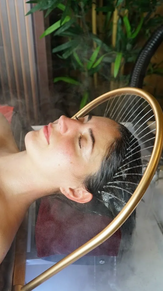 Woman lying with head in a steam facial device, surrounded by green plants.