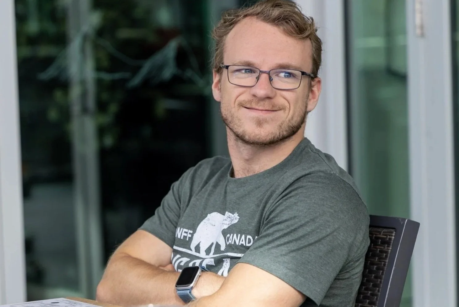 A young man with glasses and a beard sitting with arms crossed, smiling, wearing a green T-shirt and smartwatch, outdoors with glass windows in the background.