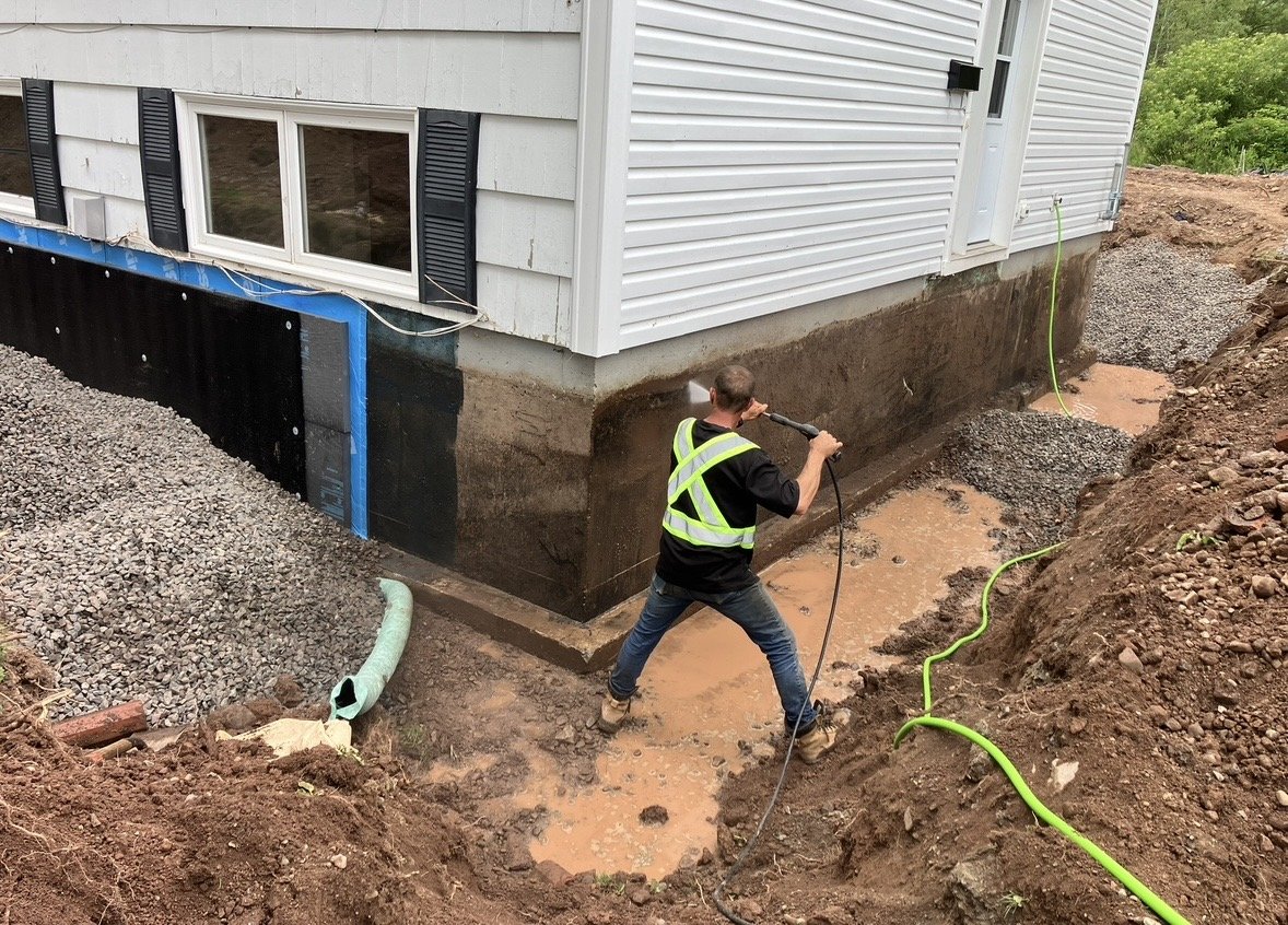 A person wearing a safety vest power washing the foundation of a house outside in a muddy area.