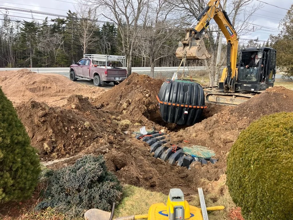 Construction site with a compact excavator lifting a large black septic tank into an underground installation, surrounded by dirt mounds, a pickup truck nearby, and some green shrubs and trees.