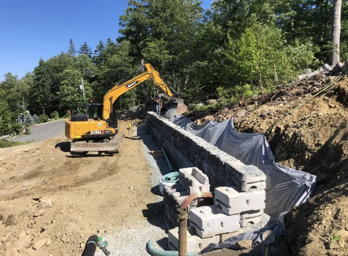 Construction site with a yellow excavator building a stone wall along a slope, with trees and a clear blue sky in the background.