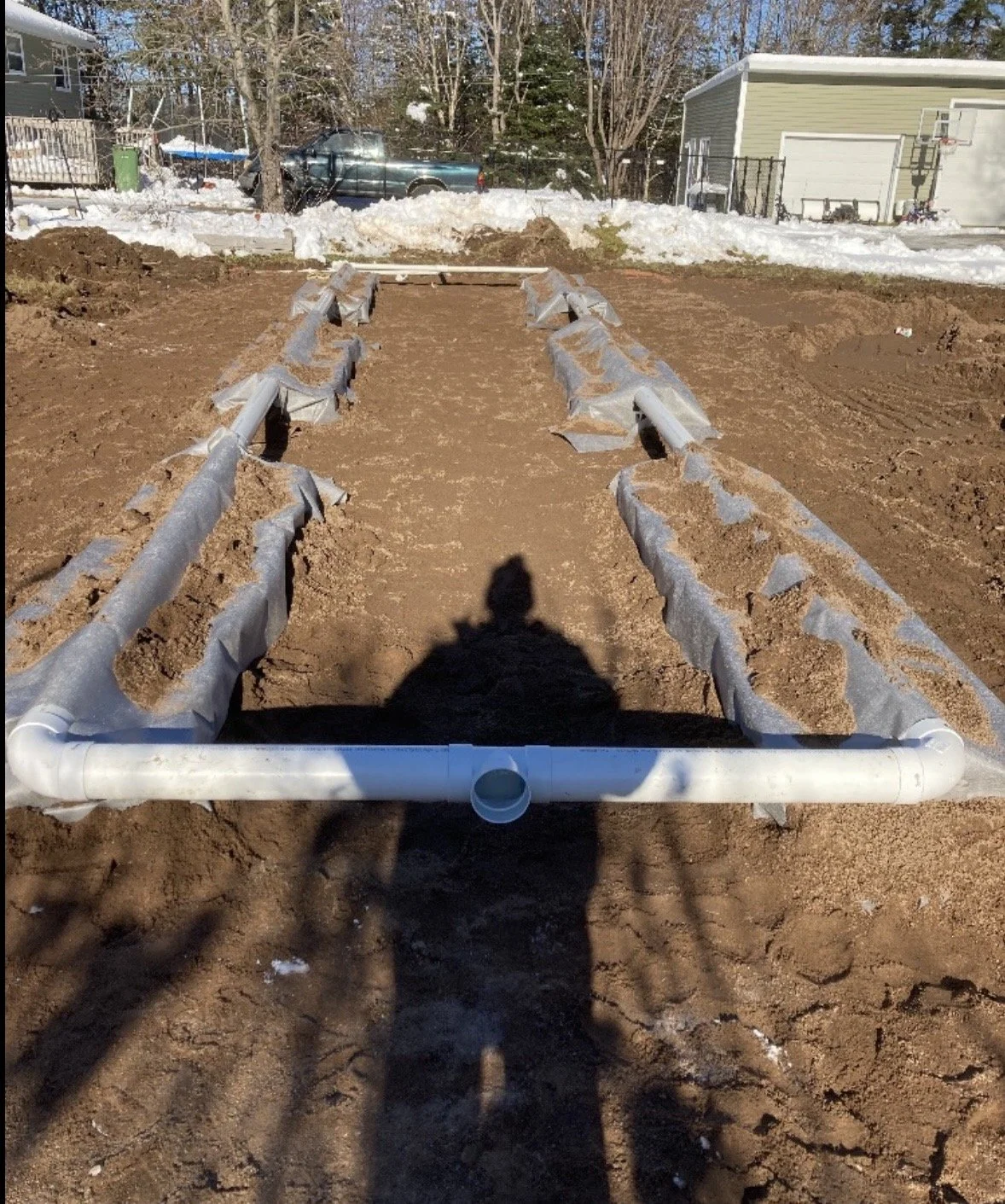 A garden bed with wooden edges and a PVC pipe rectangle frame, set in brown soil with some snow and backyard trees and houses in the background.