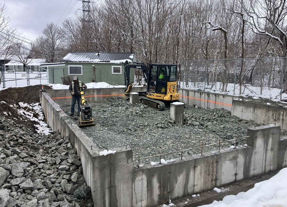 Construction site with two workers and a small excavator working on a gravel base, surrounded by snow and a chain-link fence.