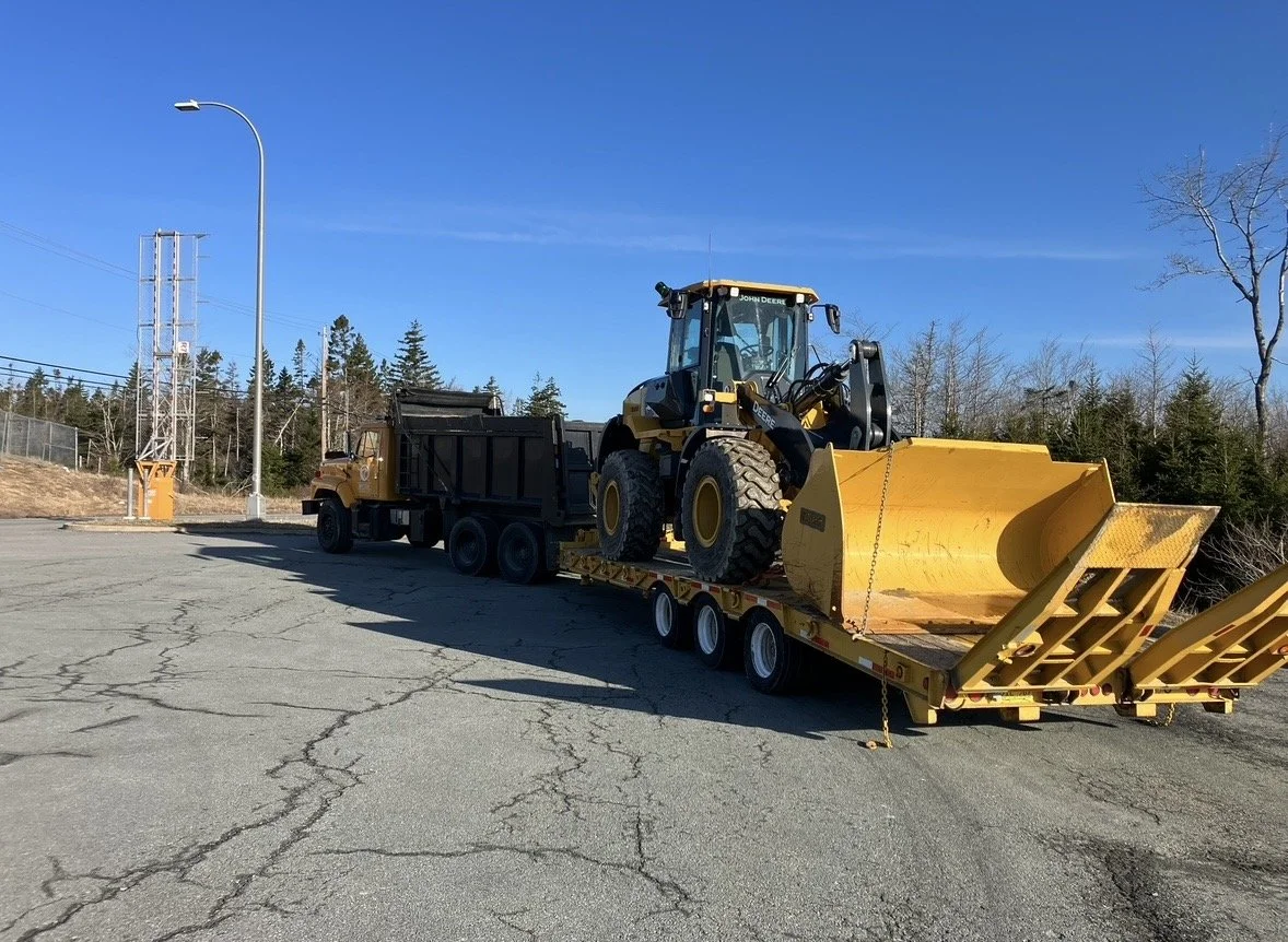 A large John Deere bulldozer loaded on a flatbed trailer, parked in a cracked asphalt lot with trees and power lines in the background.