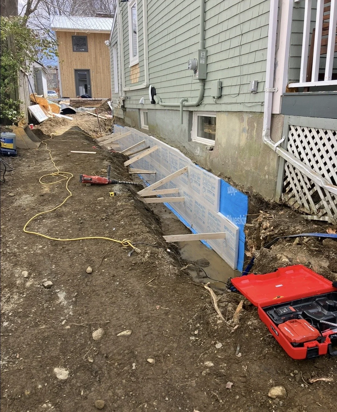 Construction work on the exterior of a house, with a trench dug along the foundation, wooden supports, insulation panels, construction tools, and a red toolbox.