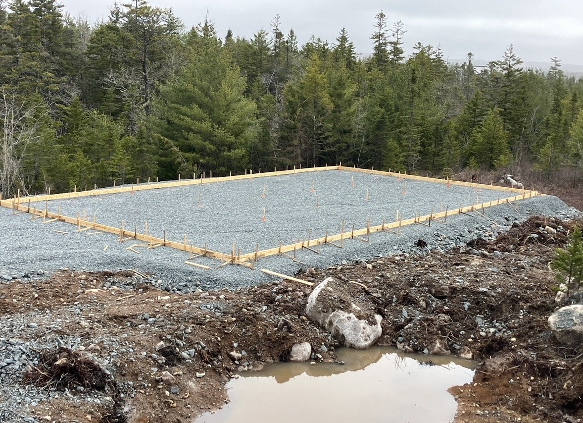 Construction site for a building foundation in a forested area, with gravel and wooden formwork in place.