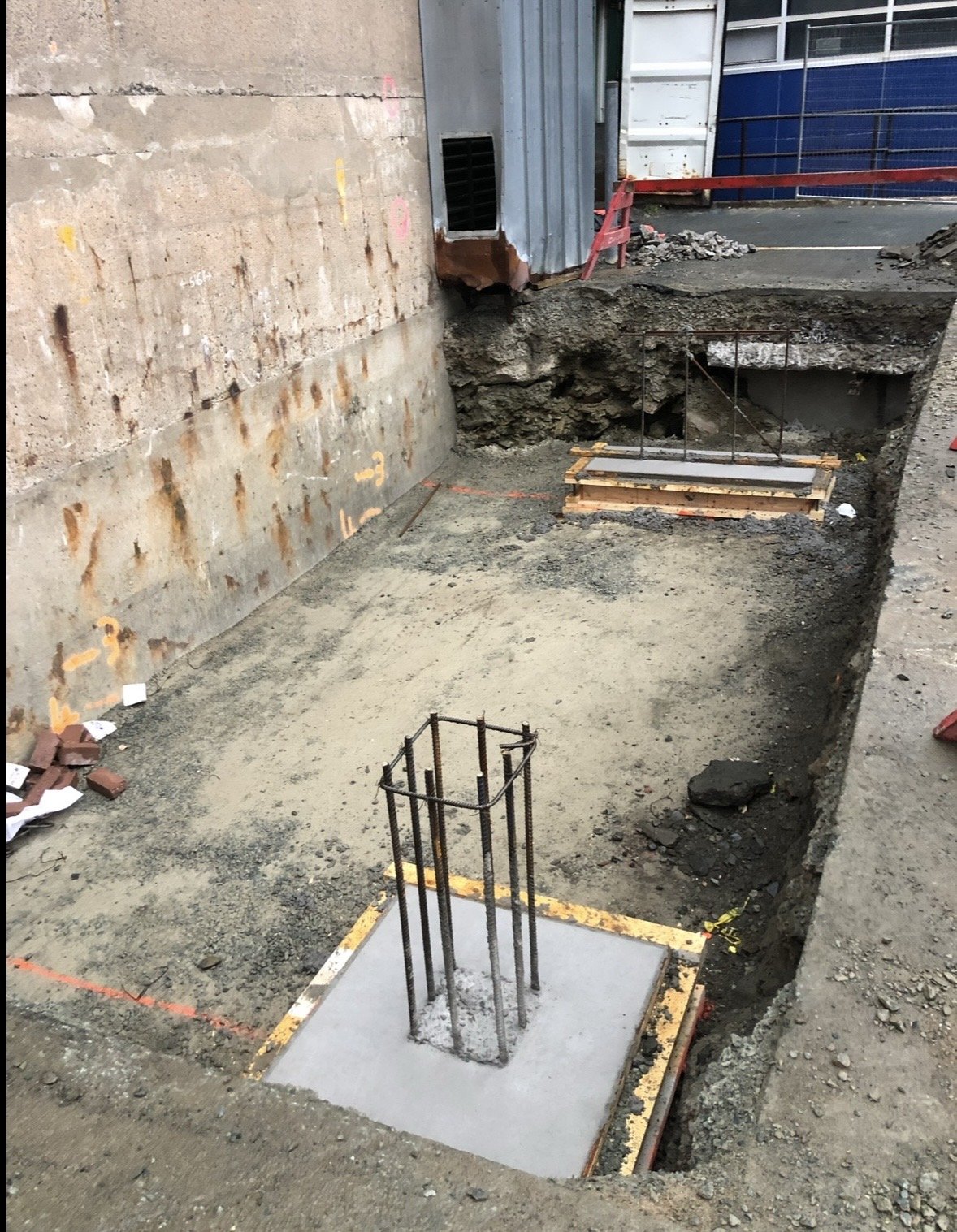 Excavation site with foundation rebar and concrete for a new structure, surrounded by dirt and construction barriers.