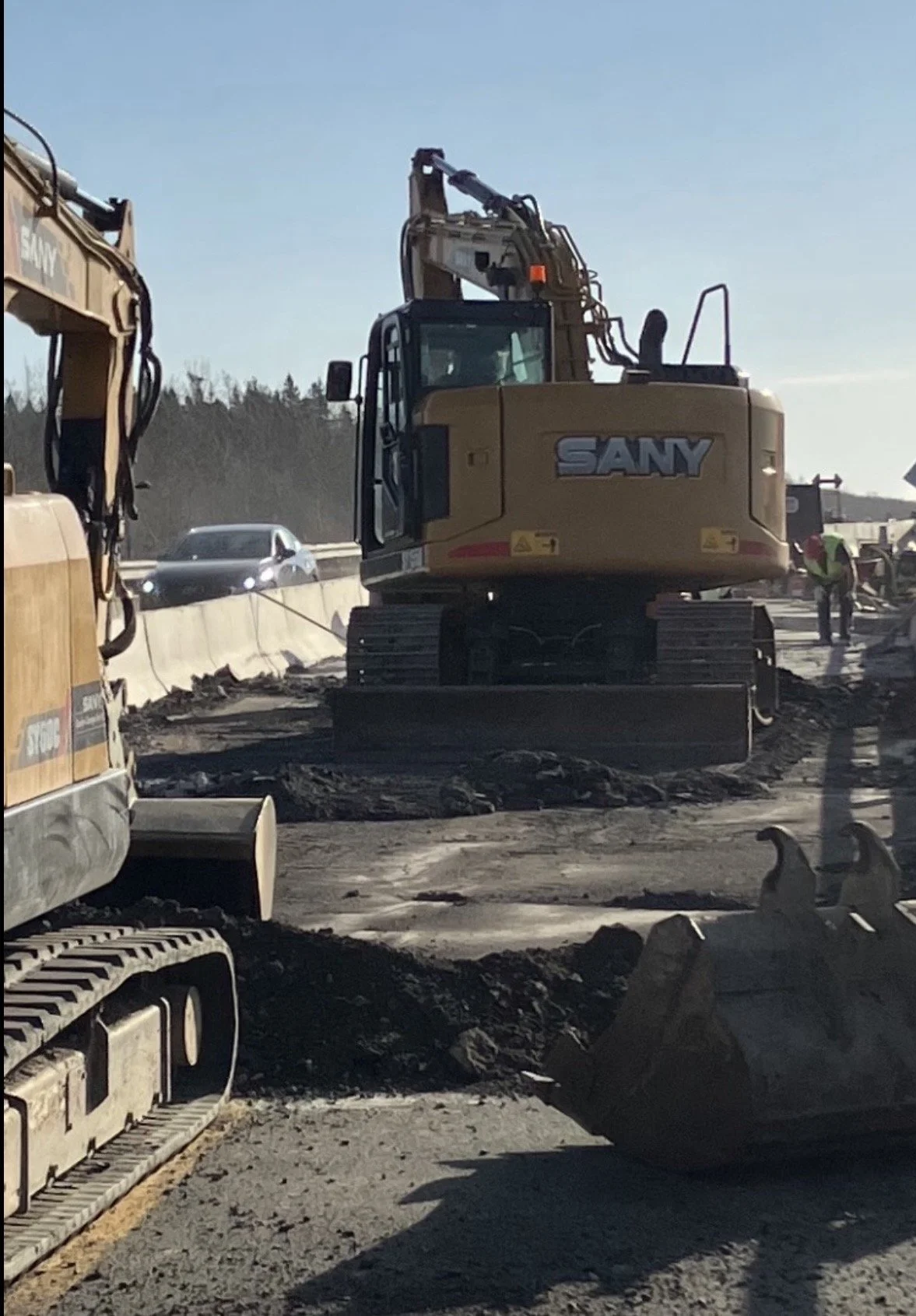 Construction site with heavy machinery, including a SANY excavator, working on a road as cars pass by on the highway in the background.