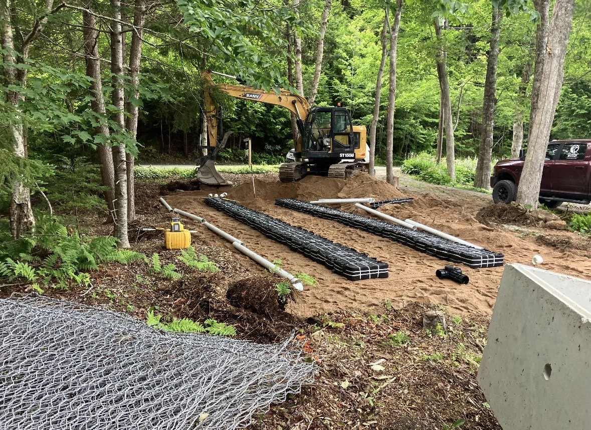 Construction site in a wooded area with a small excavator, concrete blocks, pipes, and metal grid for a project.