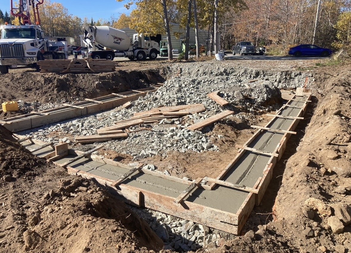 Construction site with a concrete foundation being poured in a rectangular frame, surrounded by dirt and gravel, with construction vehicles and cars parked in the background.
