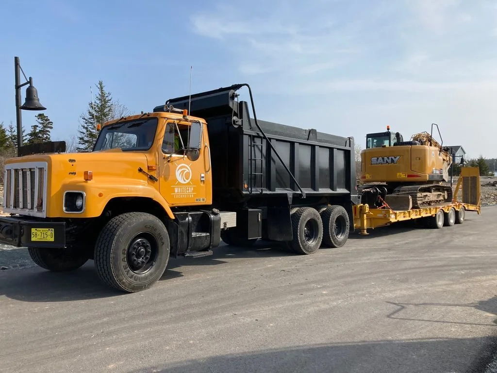 Yellow flatbed truck transporting a yellow SANY excavator on a newly paved road with a clear sky and trees in the background.