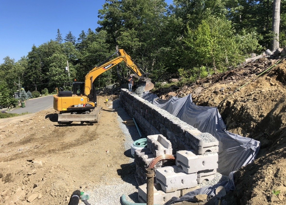 Construction site with an excavator building a stone wall, surrounded by dirt, gravel, and greenery.