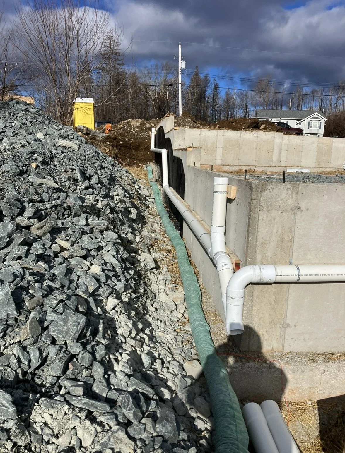 Construction site with pipes installed along a concrete foundation, large pile of rocks on the left, and dirt with a yellow portable toilet in the background.