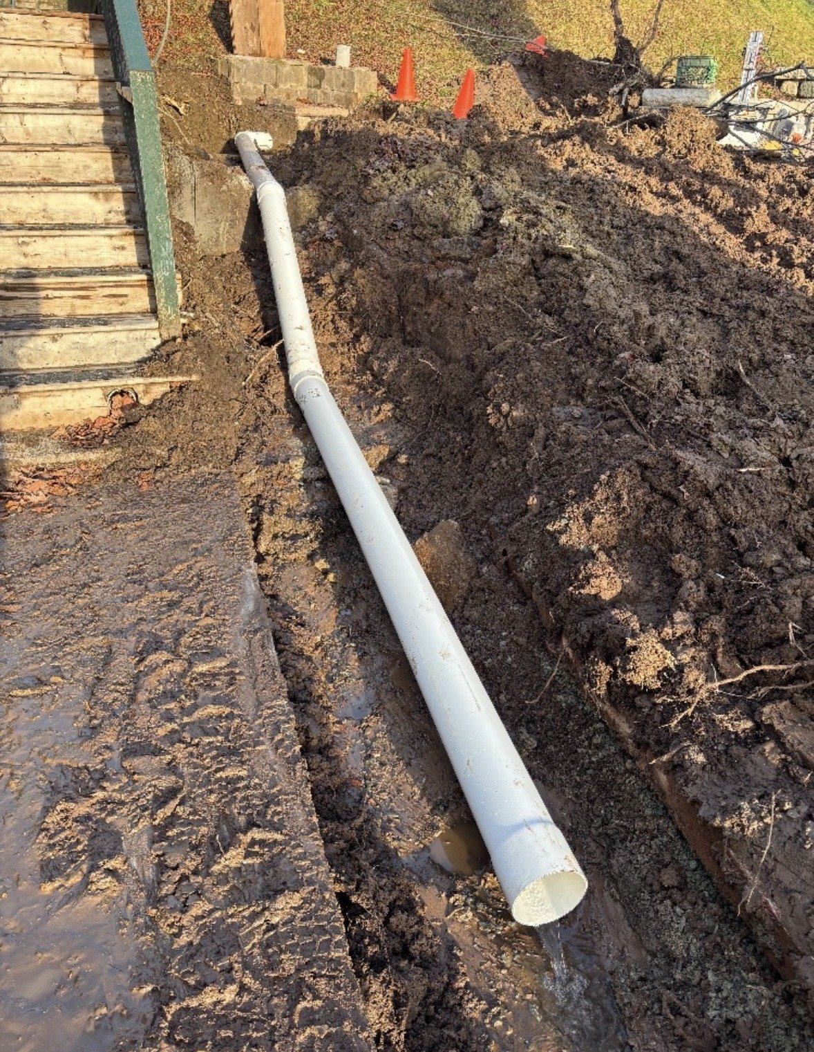 A white PVC pipe lying in a trench beside a wooden staircase, with orange construction cones and dirt surrounding it.