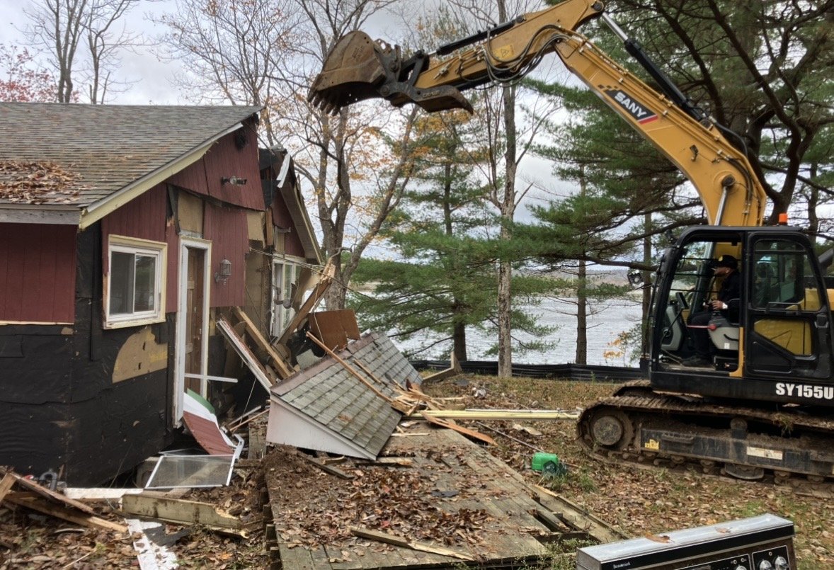 A yellow excavator demolishing a damaged house with the lake and trees in the background.