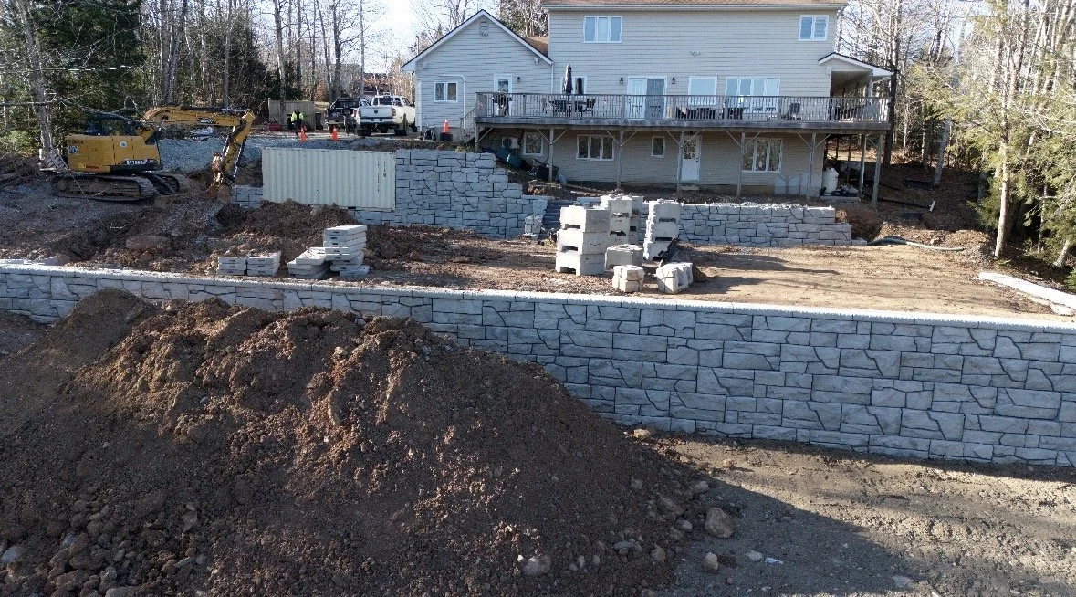 Construction site with stone retaining walls, dirt piles, and building materials in front of a house with a deck.