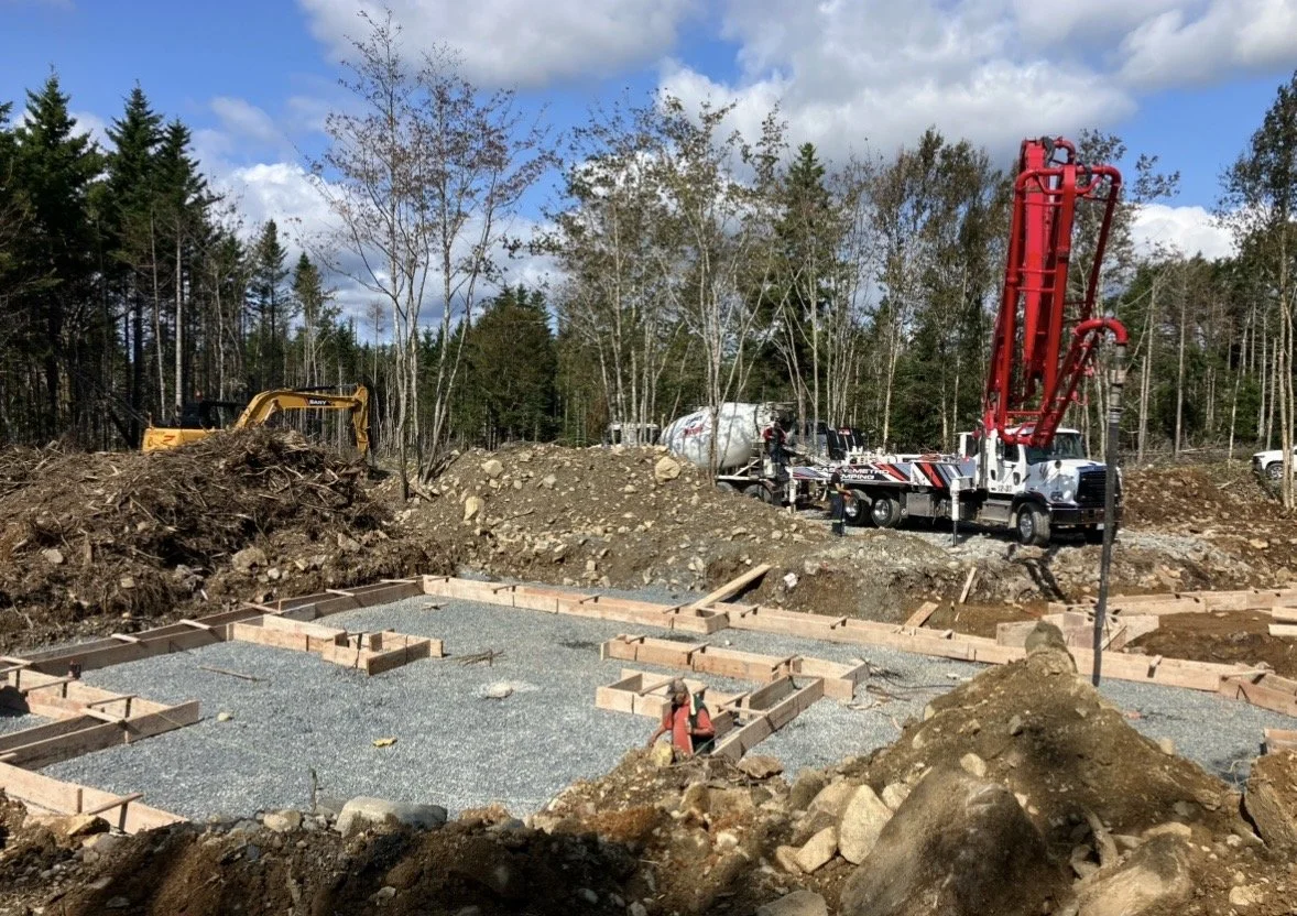 Construction site showing the foundation of a building being prepared with wooden forms and gravel. Construction machinery and trucks are present, with a backdrop of trees and a partly cloudy sky.