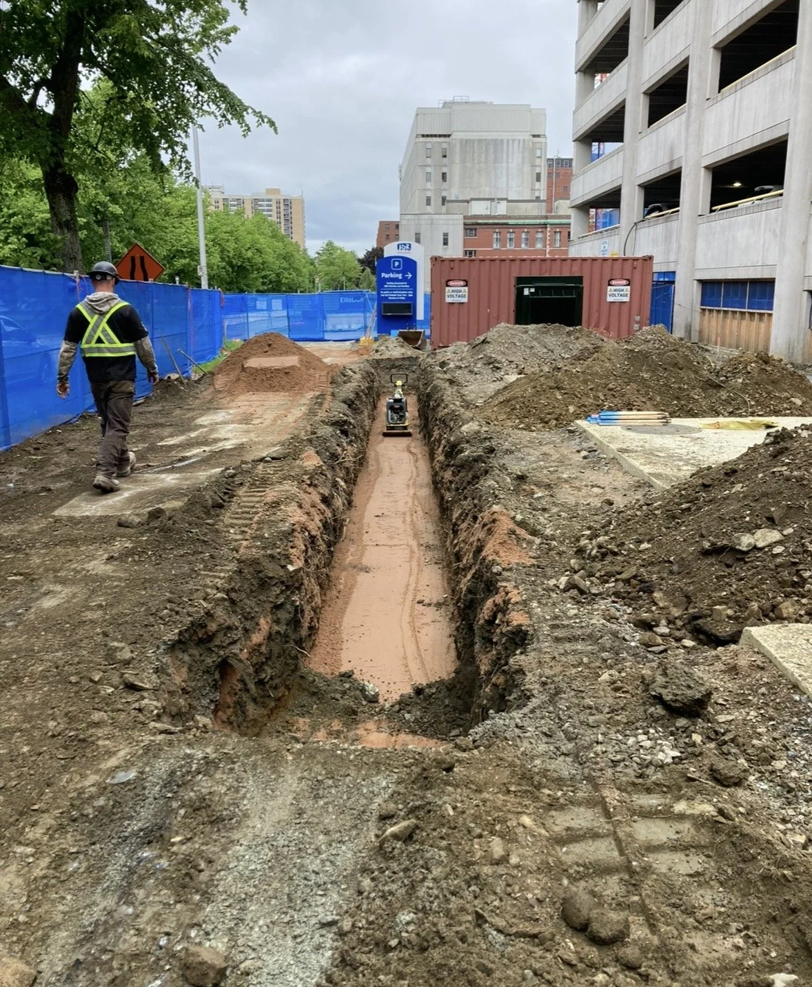 Construction site with a trench dug for utilities, a worker in safety gear walking, and construction materials around. Blue fencing and a parking lot in the background.