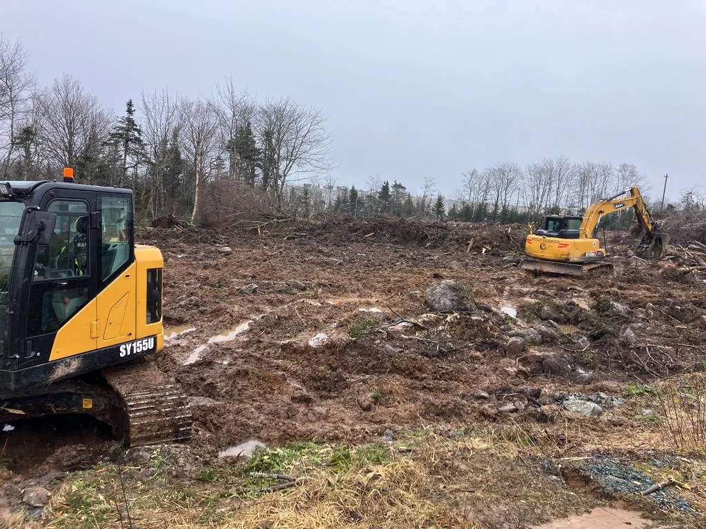 Construction site with two yellow excavators working on cleared land with rocks and dirt, trees in the background.