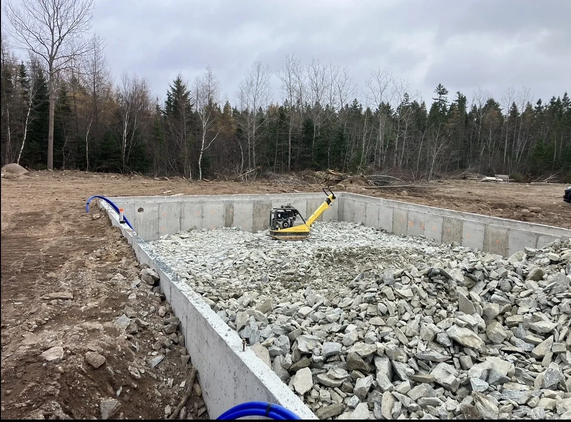 A construction site with a concrete foundation filled with gravel and a plate compactor in the center, surrounded by dirt and piled stones, with a backdrop of leafless trees and cloudy sky.