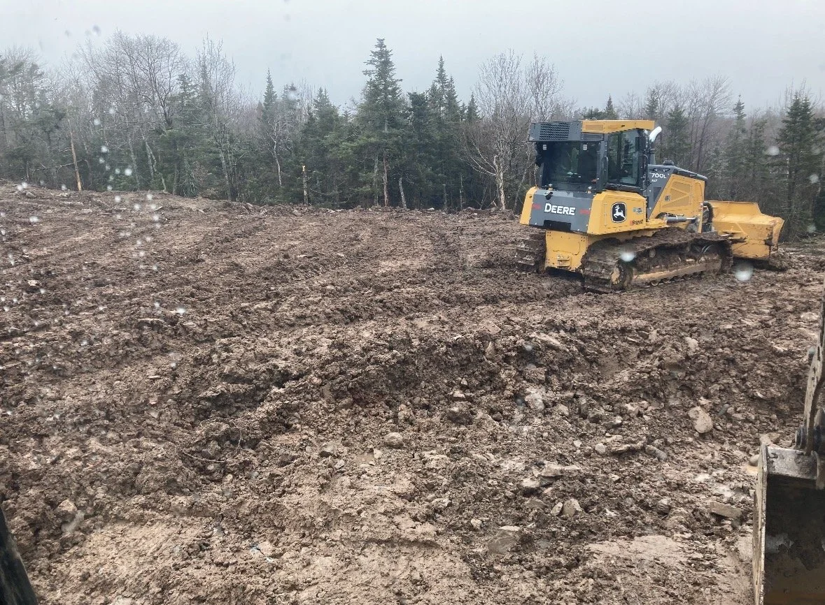 A John Deere bulldozer working on a muddy construction site with trees in the background.