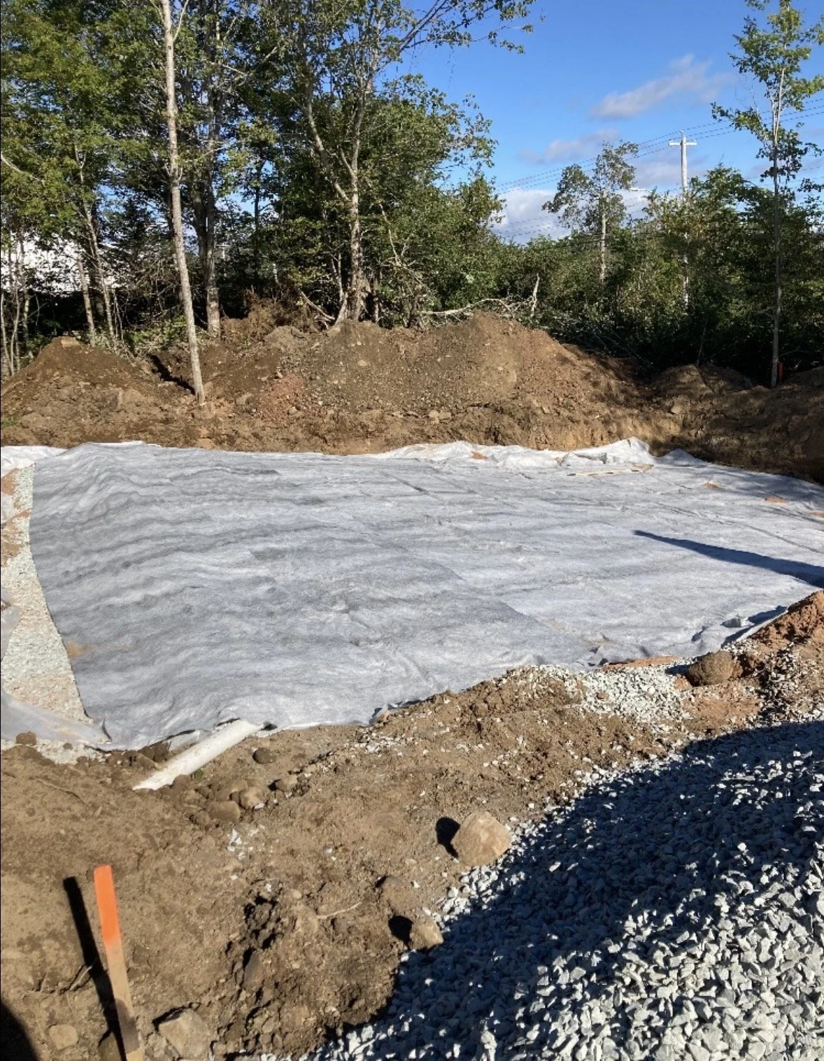 Construction site with a foundation covered by a white vapor barrier, surrounded by dirt and gravel, and trees in the background under a blue sky with clouds.