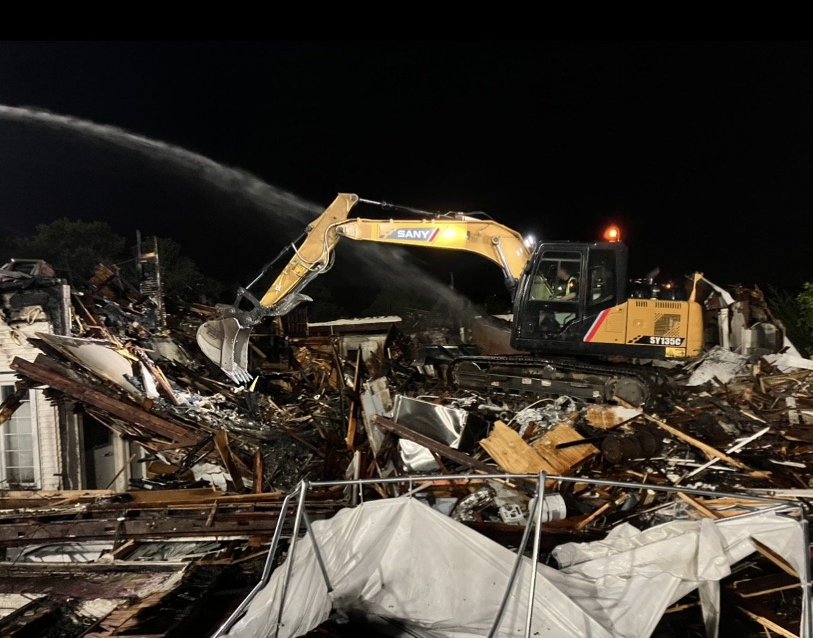 A construction excavator operates amidst the wreckage of a destroyed building at night.