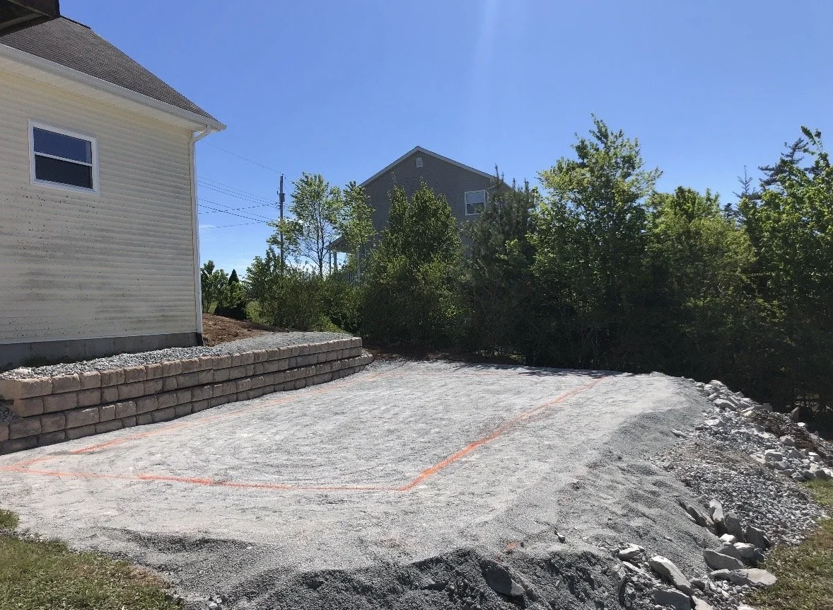 A backyard with a gravel foundation marked for construction, a small retaining wall made of bricks, and a house on the left with lush green trees and another house in the background under a clear blue sky.