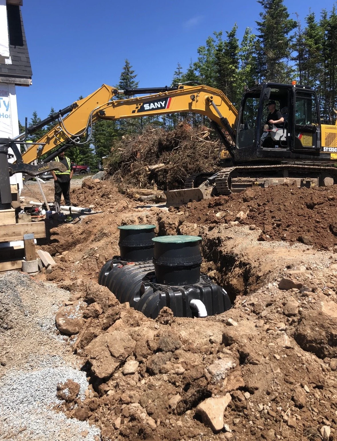 A construction site with a yellow excavator digging earth near underground drainage tanks, with workers and trees in the background.