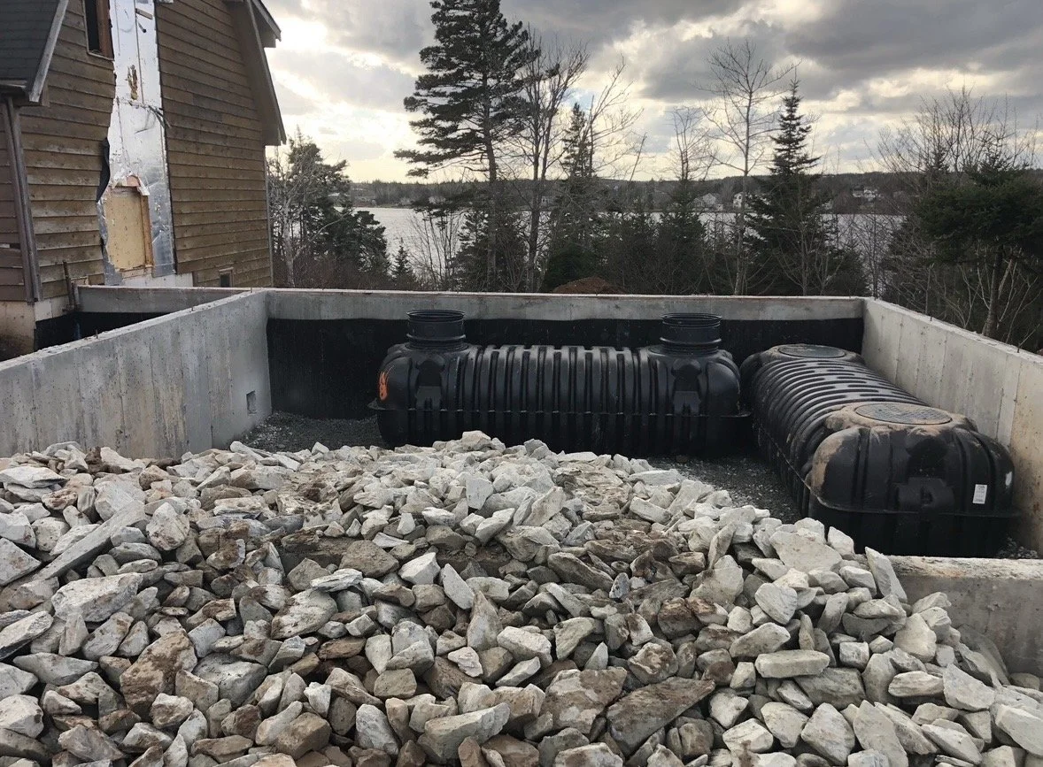 Construction site with two black septic tanks installed in a concrete basin, surrounded by white and gray rocks, with a wooded landscape and lake in the background under cloudy skies.