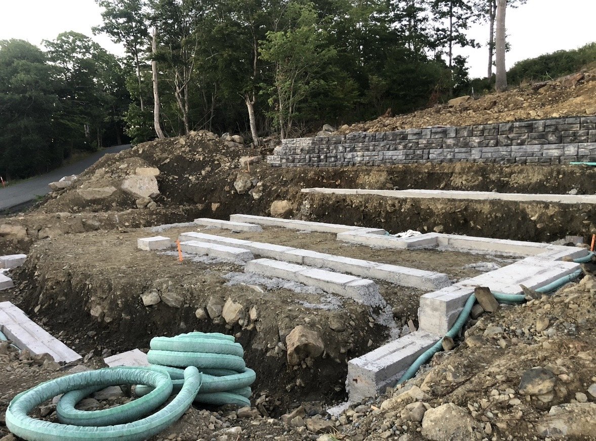 Construction site with concrete foundation and green flexible tubing, trees and rocky terrain in the background.