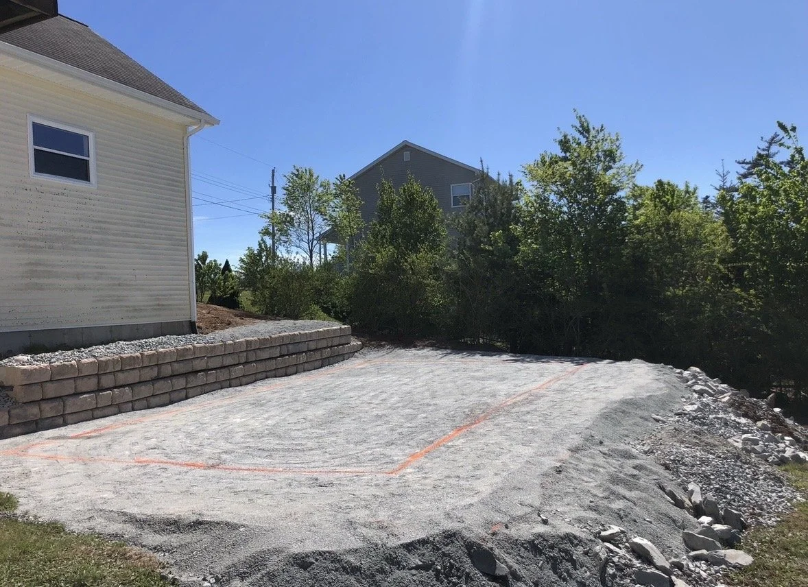 A cleared and leveled residential driveway or parking area under construction, with orange markings and a stone retaining wall next to a yellow house, surrounded by trees and a clear blue sky.
