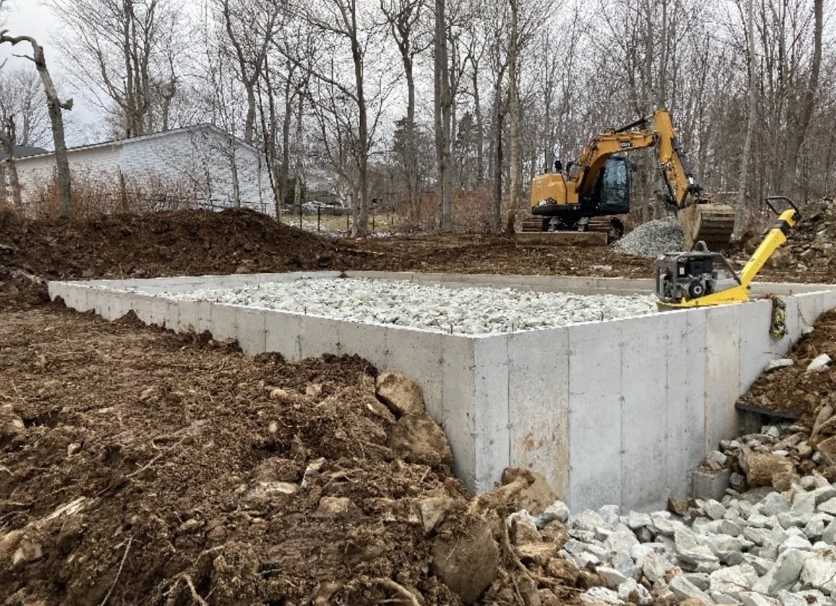 Construction site with a concrete foundation, a yellow excavator, and a compacting machine, in a wooded area.