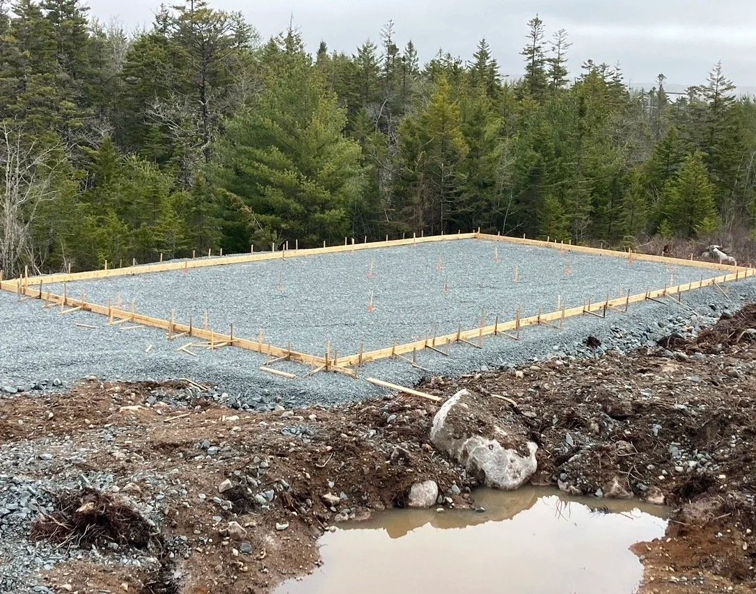 A construction site for a building foundation with gravel, surrounded by a wooded area.